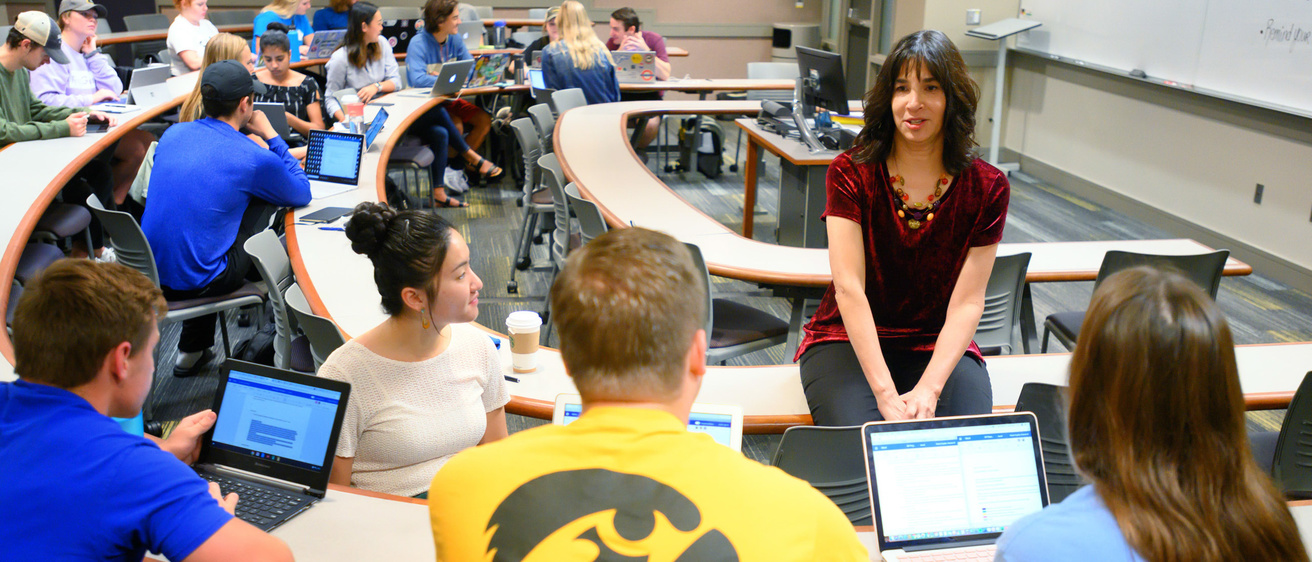 alt="Professor sitting on desk teaching to four seated students."