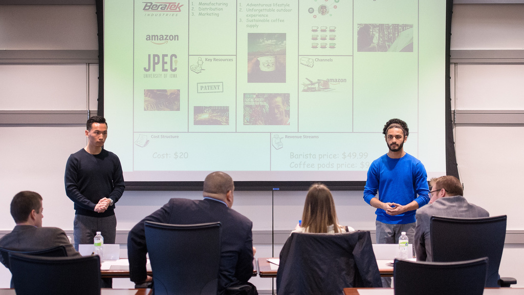 Two students presenting their business plan stand in front of four seated people.